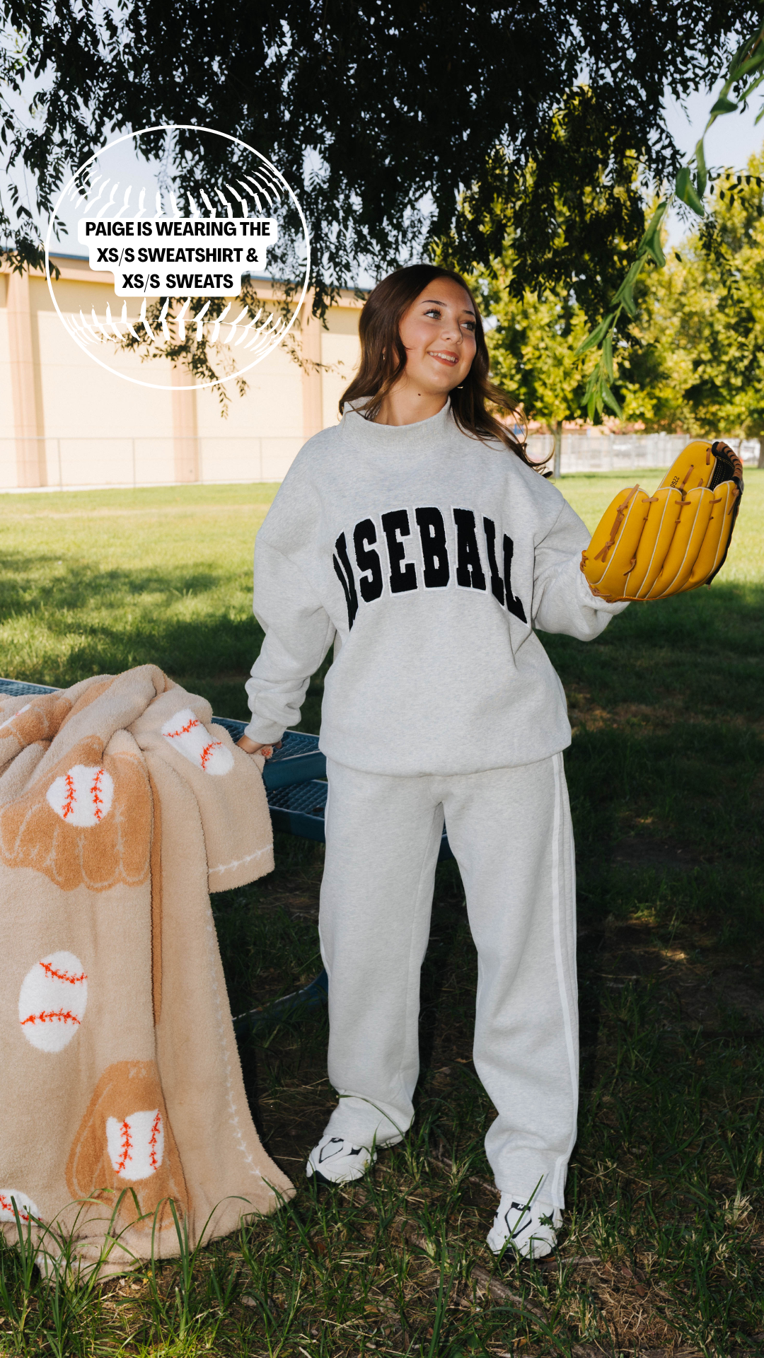 A woman standing outdoors on grass wearing a light gray sweatshirt and sweatpants set with 'BASEBALL' printed in black across the chest. She is holding a yellow baseball glove and a tan blanket with baseball patterns.