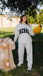 A woman standing outdoors on grass wearing a light gray sweatshirt and sweatpants set with 'BASEBALL' printed in black across the chest. She is holding a yellow baseball glove and a tan blanket with baseball patterns.