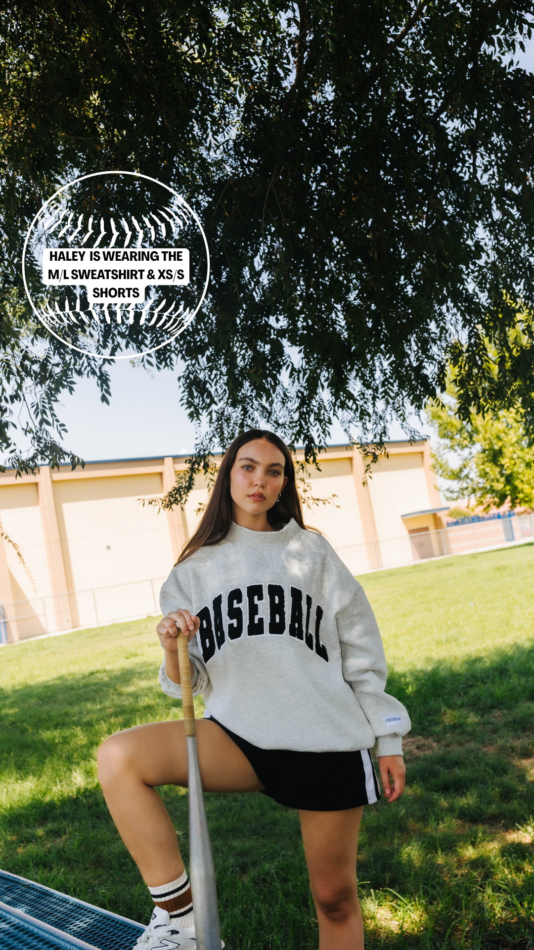 A woman outdoors on a grassy field under a tree, wearing a white sweatshirt with 'BASEBALL' printed in black across the chest, black shorts, and white sneakers.