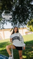 A woman outdoors on a grassy field under a tree, wearing a white sweatshirt with 'BASEBALL' printed in black across the chest, black shorts, and white sneakers.