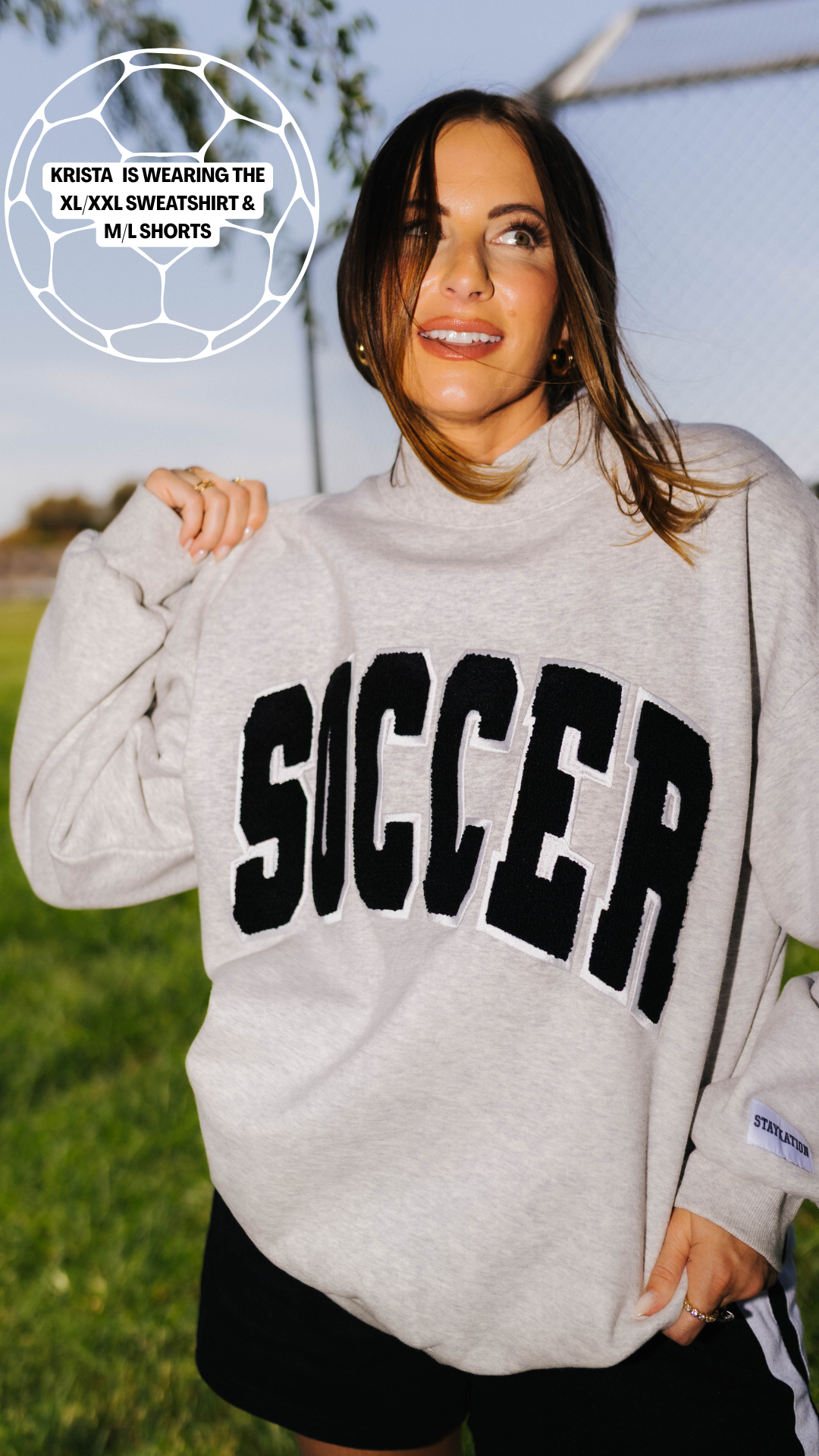 A woman outdoors wearing a light heather gray sweatshirt with the word 'SOCCER' in large black varsity-style letters outlined in white. She is also wearing black athletic shorts, with a grassy field and sky in the background.