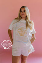 Woman wearing a white t-shirt with a shell graphic and shorts on a pink background size 2xl