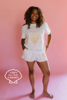 Woman wearing a white t-shirt with a shell design and shorts on a pink background size L/S