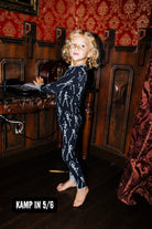 A young child wearing a black long-sleeve pajama set with a white skeleton pattern, standing barefoot in a dark wood room with red patterned wallpaper. A text overlay at the bottom left reads 'KAMP IN 5/6'.