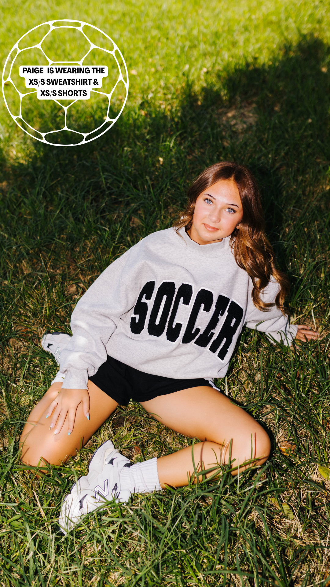 A woman lying on green grass wearing a light gray sweatshirt with 'SOCCER' printed in black across the chest, black shorts, white socks, and white sneakers. A white soccer ball outline graphic with text is in the top-left corner.