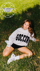 A woman lying on green grass wearing a light gray sweatshirt with 'SOCCER' printed in black across the chest, black shorts, white socks, and white sneakers. A white soccer ball outline graphic with text is in the top-left corner.