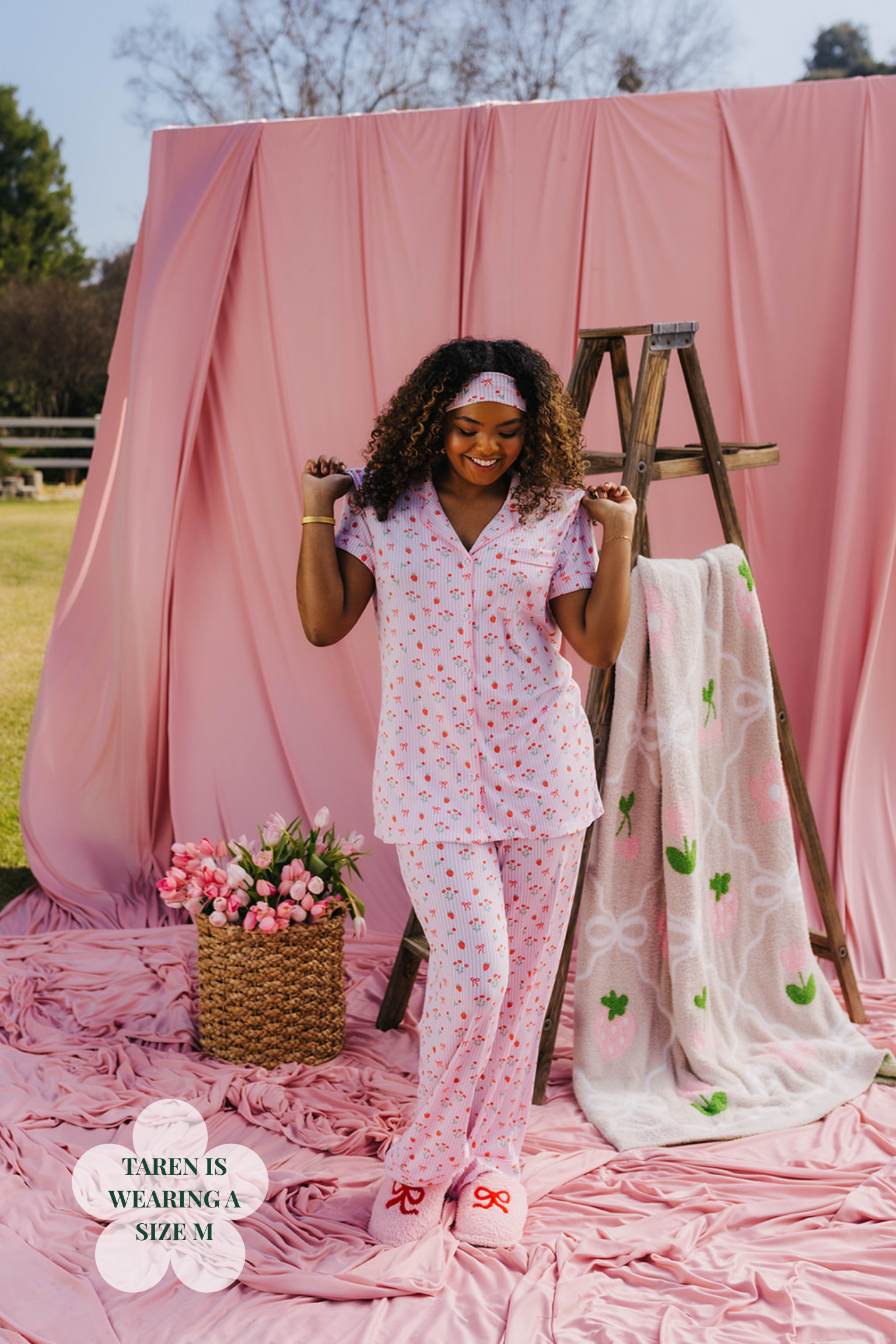 Woman in a pink floral pajama set standing in front of a pink backdrop with a ladder and flowers.