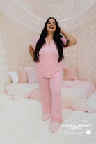 Woman wearing pink pajamas in a cozy bedroom setting with fairy lights.