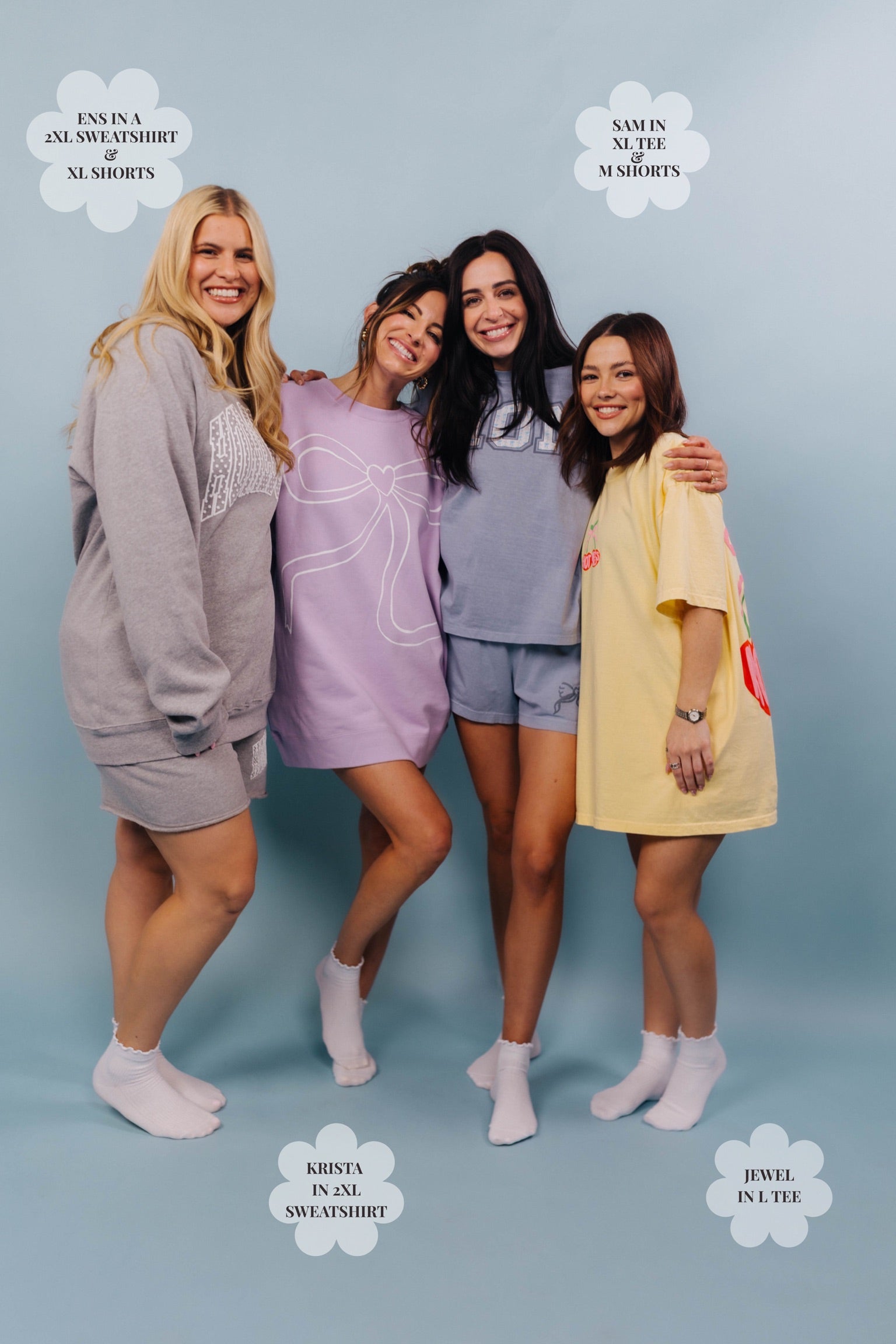 Four women posing together wearing different styles of shorts and merch on a light blue background.