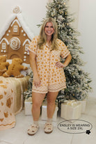 Woman in a gingerbread pajama set standing in front of a Christmas tree and gingerbread house.