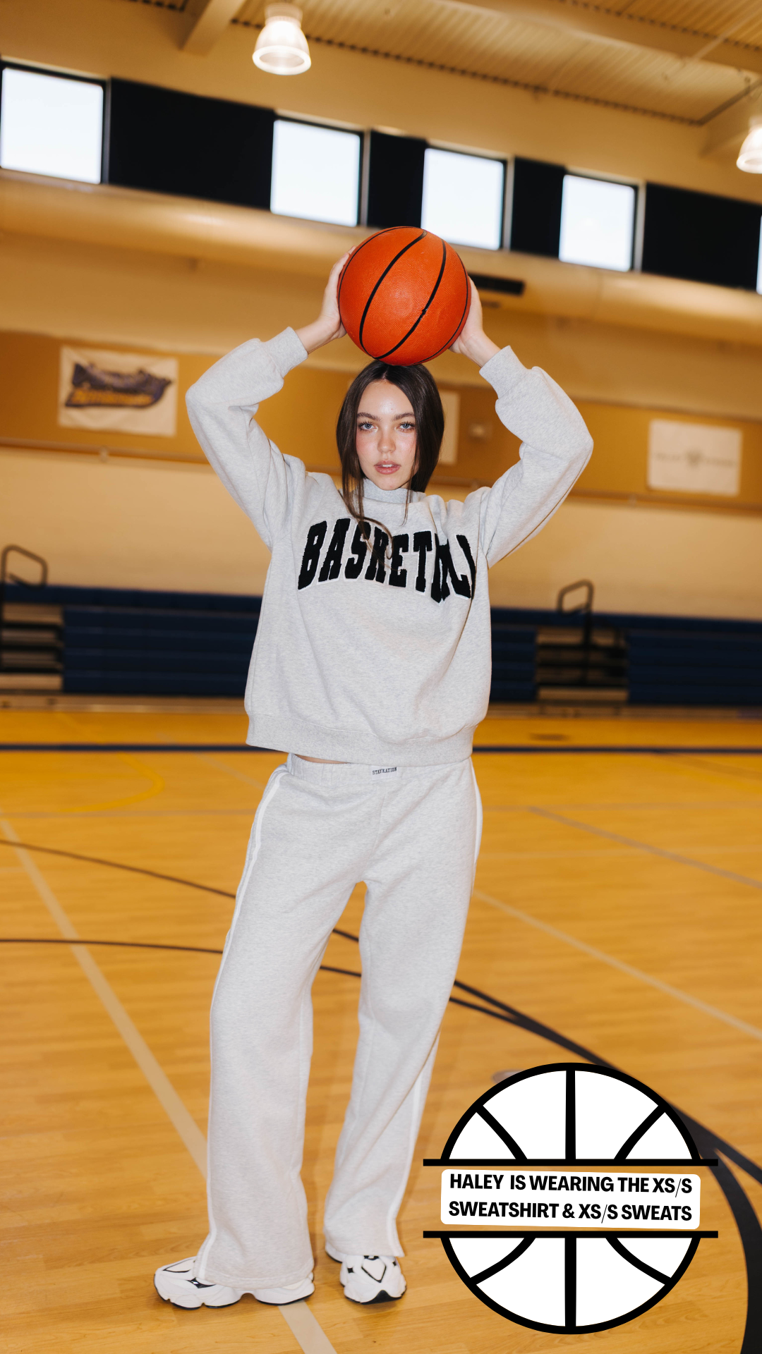 A woman stands on a basketball court wearing a light gray sweatshirt and sweatpants set with black 'BASKETBALL' lettering across the chest. She is holding an orange basketball above her head.