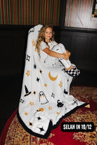 A young girl sits wrapped in a white Halloween-themed blanket featuring black and gold witch motifs, including stars, ghosts, a moon, and a witch hat, in an indoor setting with a patterned rug and dark paneled walls.