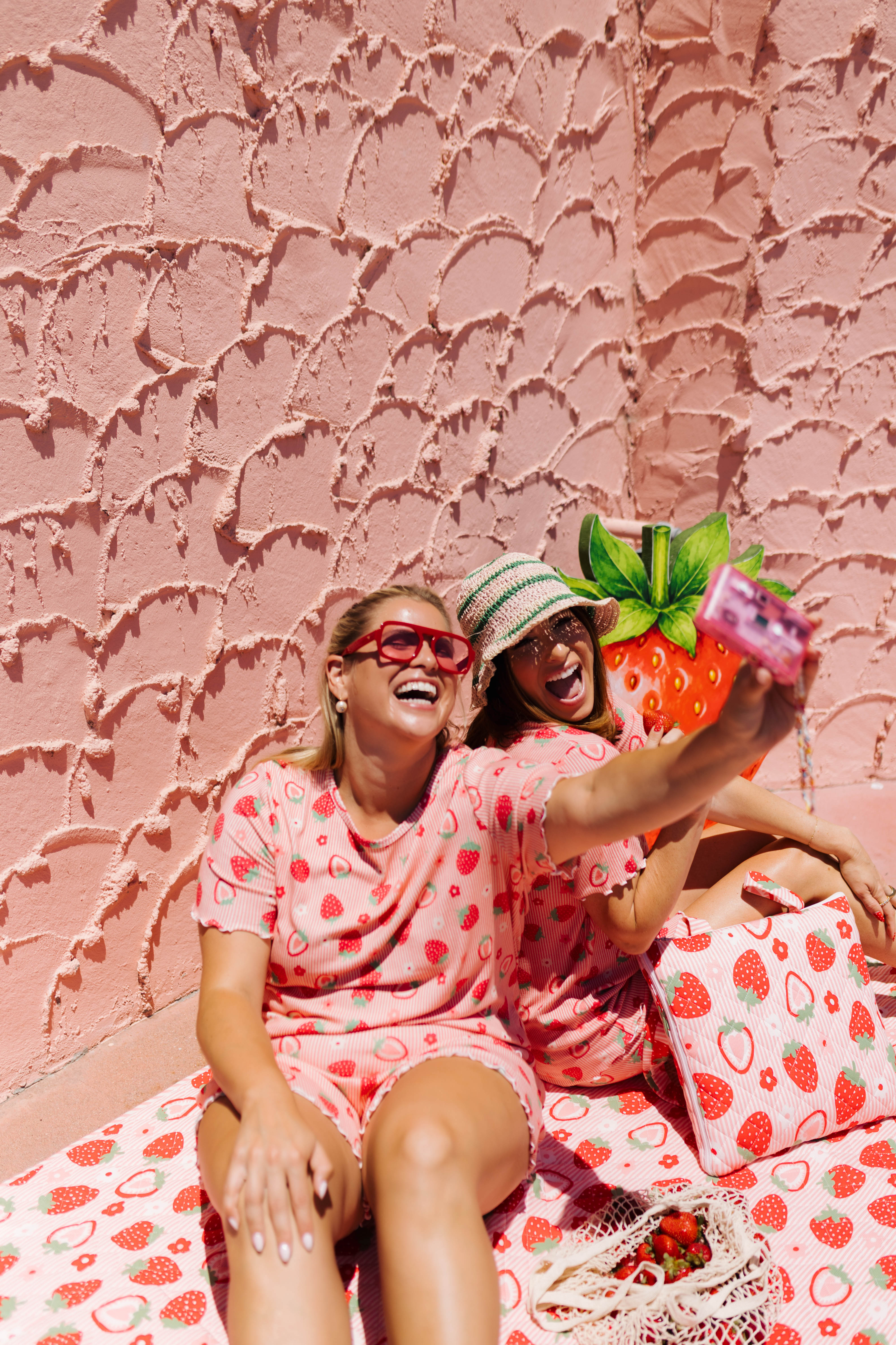 Two women wearing matching pink strawberry-print pajamas take a selfie in front of a textured pink wall. 