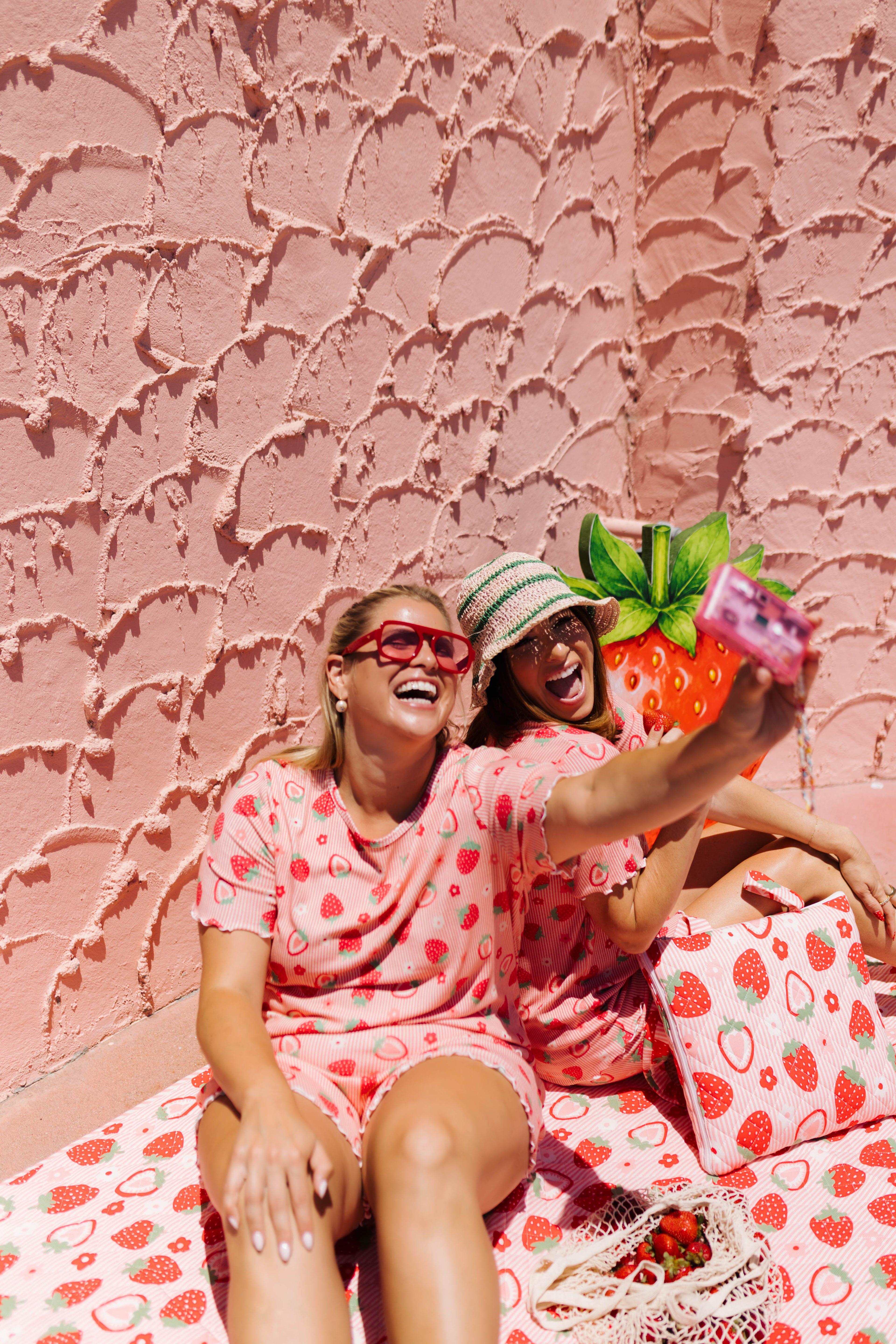 Two women wearing matching pink strawberry-print pajamas take a selfie in front of a textured pink wall. 