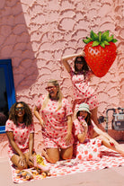Four women posing in matching pink strawberry-print pajamas in front of a pink textured building with blue window frames. One woman holds a large strawberry-shaped fruit.