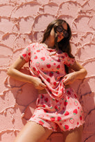 A woman wearing a pink strawberry-print short-sleeve top and matching shorts pj set posing against a textured pink wall.