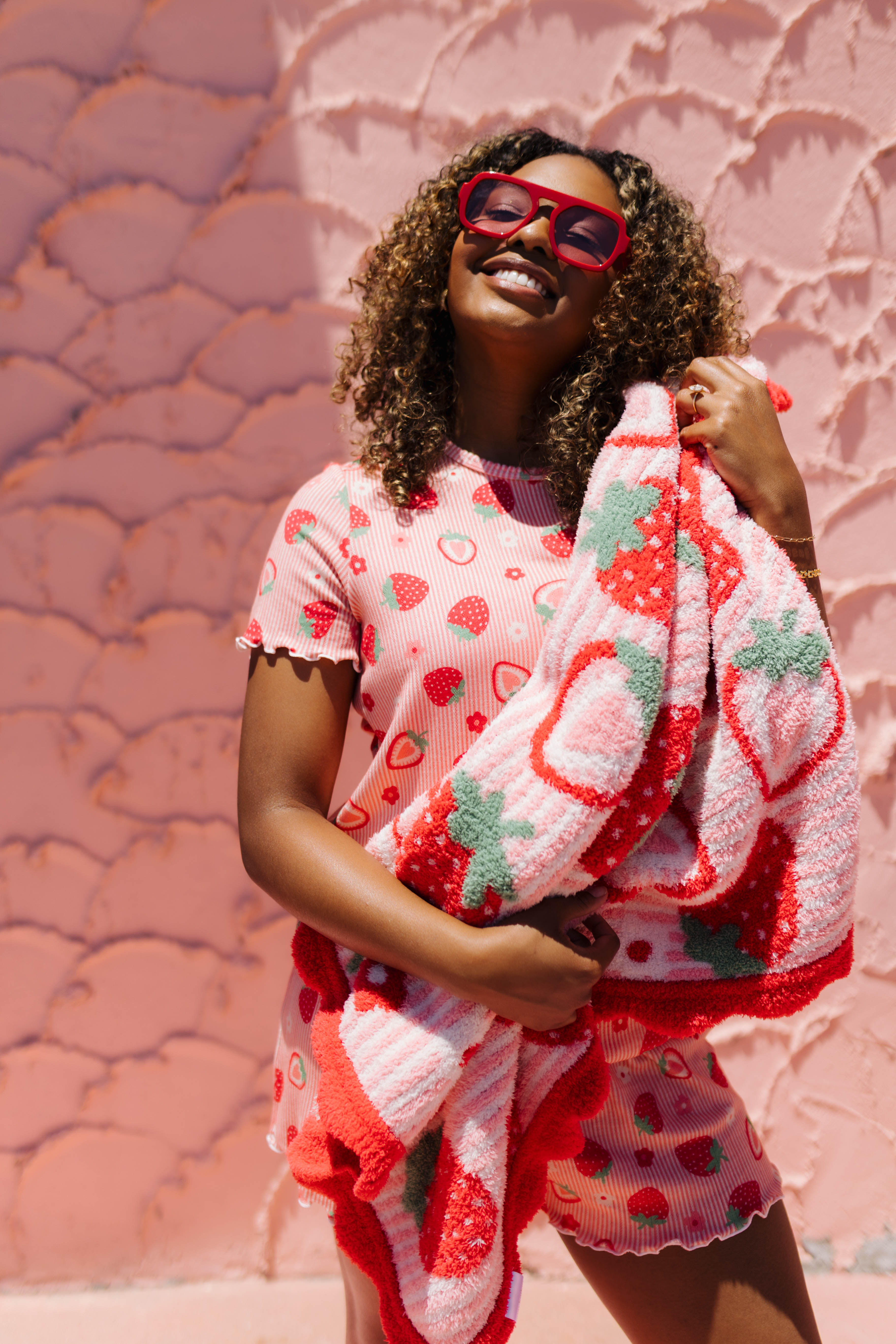 A woman wearing a pink strawberry-pj holding a matching blanket smiling and wearing red-framed sunglasses.