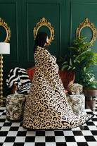 Person wrapped in a leopard print blanket in a stylish room with green walls, gold mirrors, and patterned furniture.