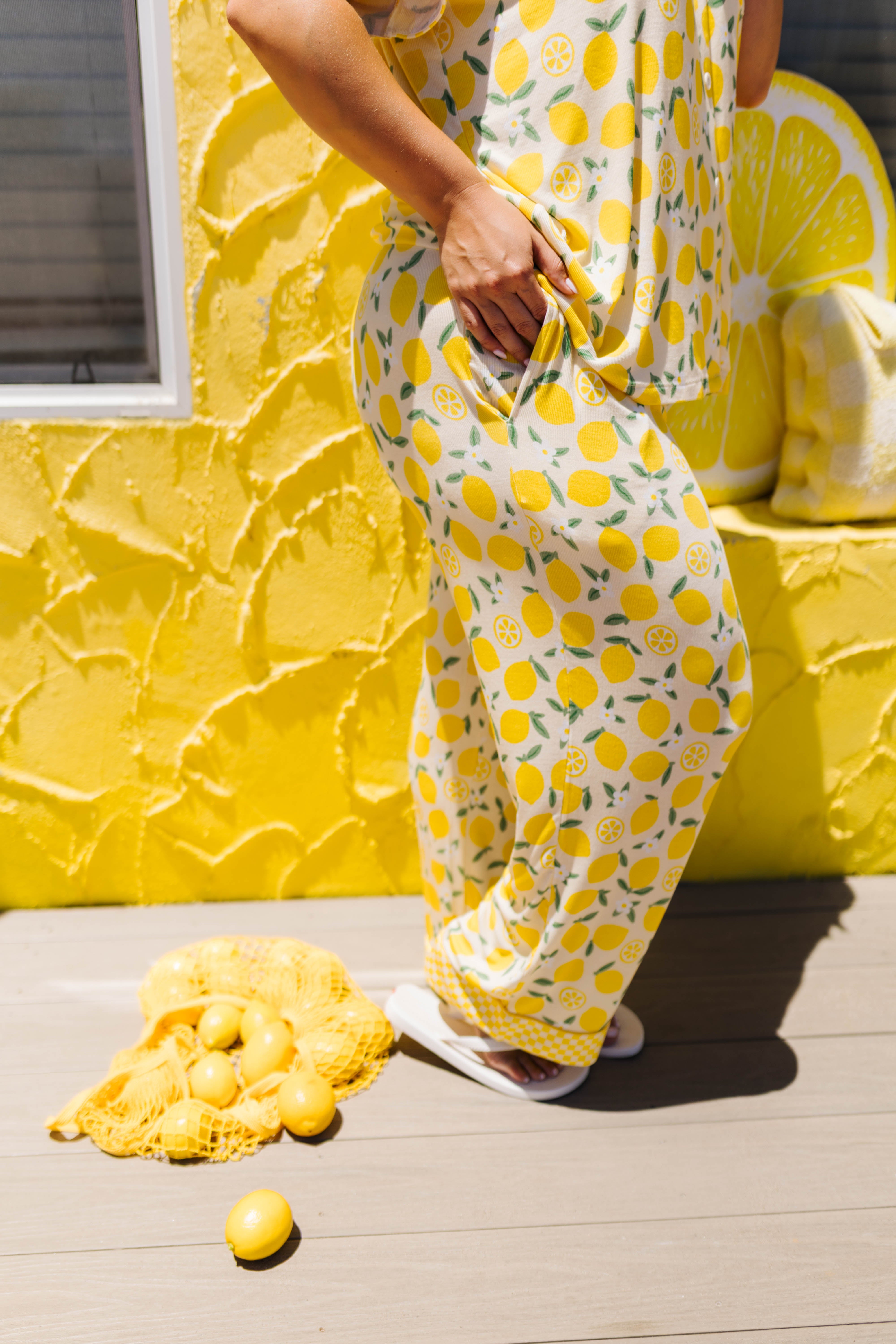 A person wearing a yellow lemon-print pj, standing on a light deck in front of a bright yellow textured wall with lemon-themed decor.