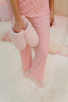 Person wearing pink polka dot pajamas holding pink slippers on a white surface.