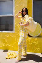 A woman wearing a yellow lemon-patterned pajama set stands in front of a bright yellow textured wall, holding a large plush quillow. A pile of lemons sits on the ground nearby.