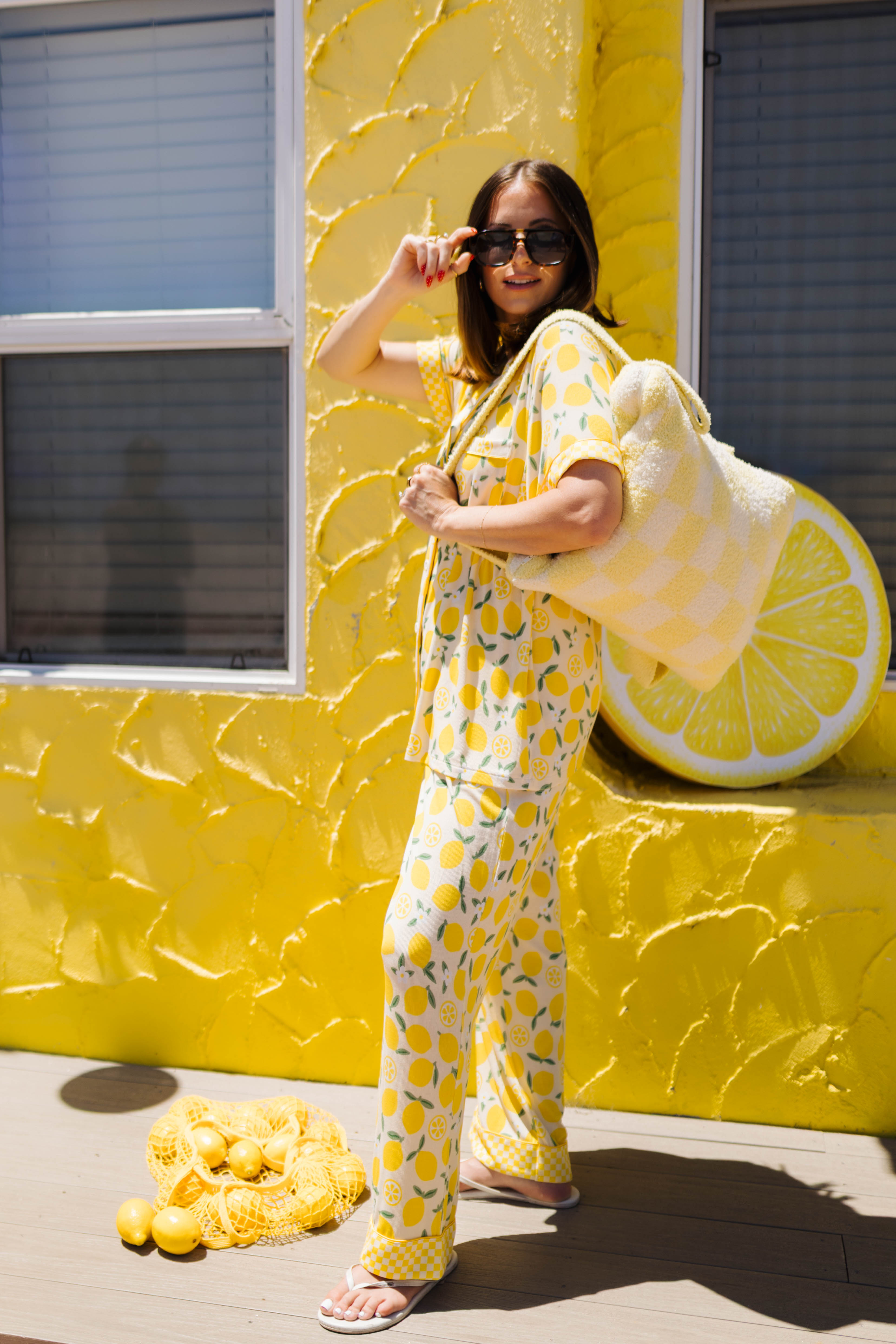 A woman wearing a yellow lemon-patterned pajama set stands in front of a bright yellow textured wall, holding a large plush quillow. A pile of lemons sits on the ground nearby.