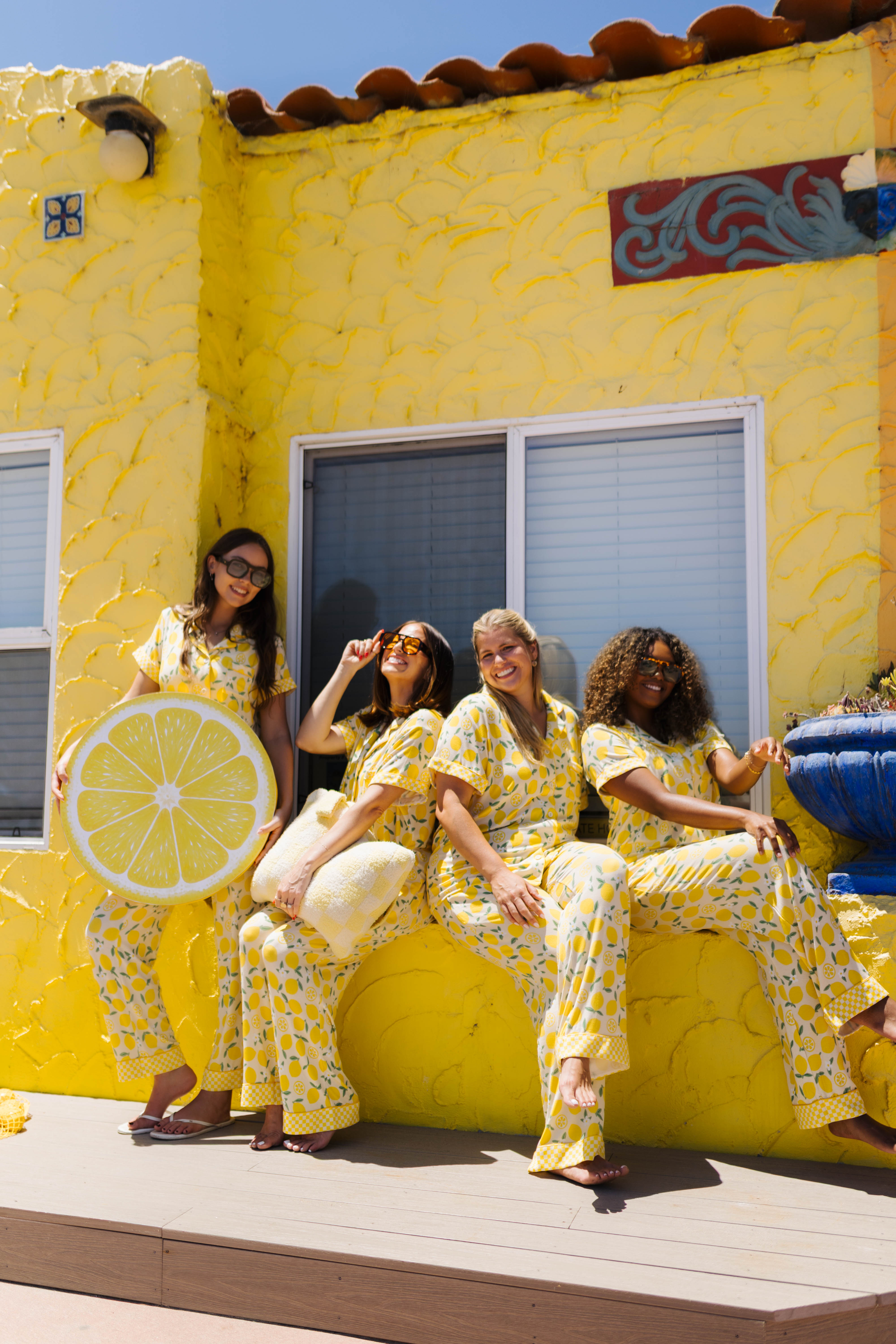 Four women wearing matching yellow and lemon-print pajamas sit on a yellow building porch, with one holding a large lemon-shape.