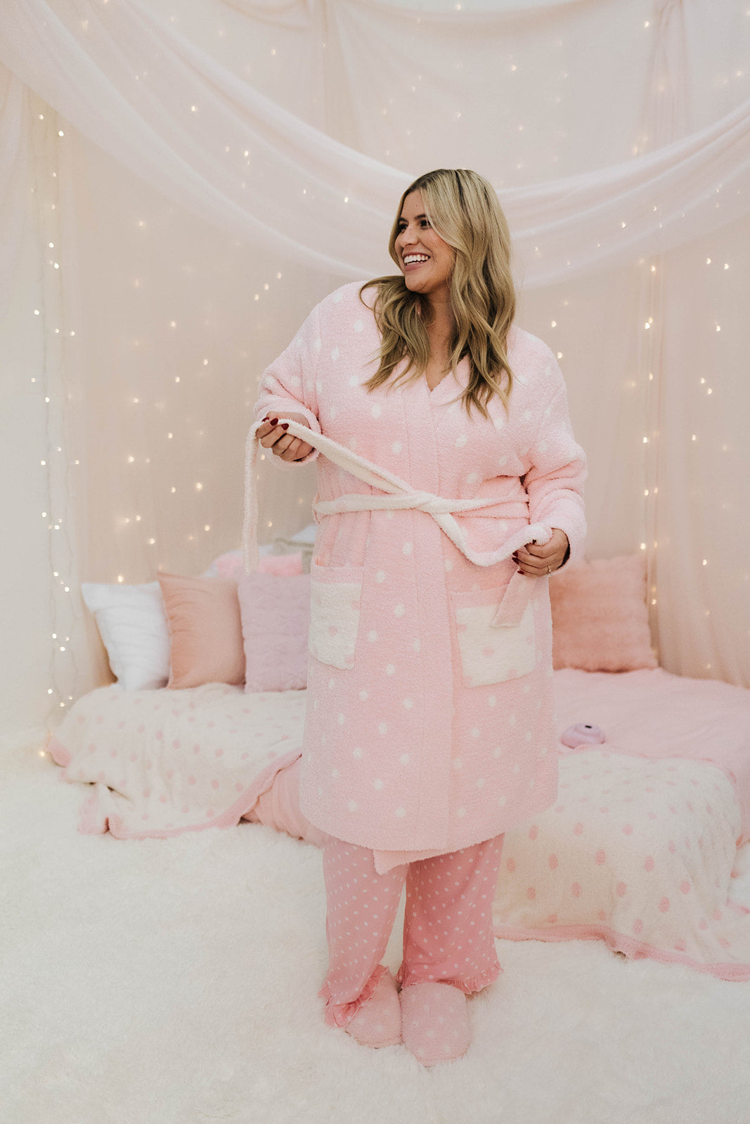 Woman wearing a pink polka dot robe in a cozy room with lights.