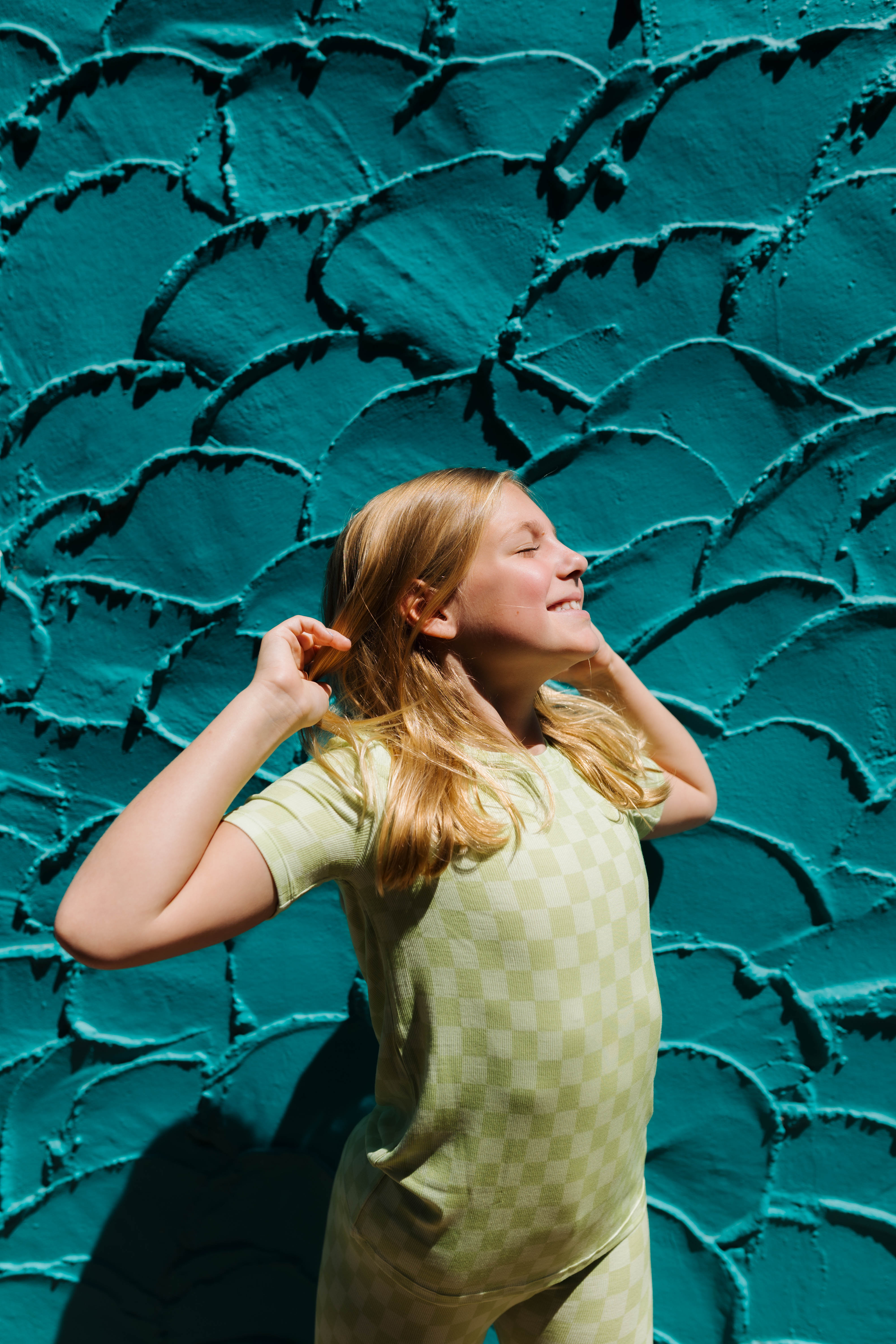 A young girl with blonde hair wearing a light green short sleeve top and matching pants, posing in front of a textured teal wall.