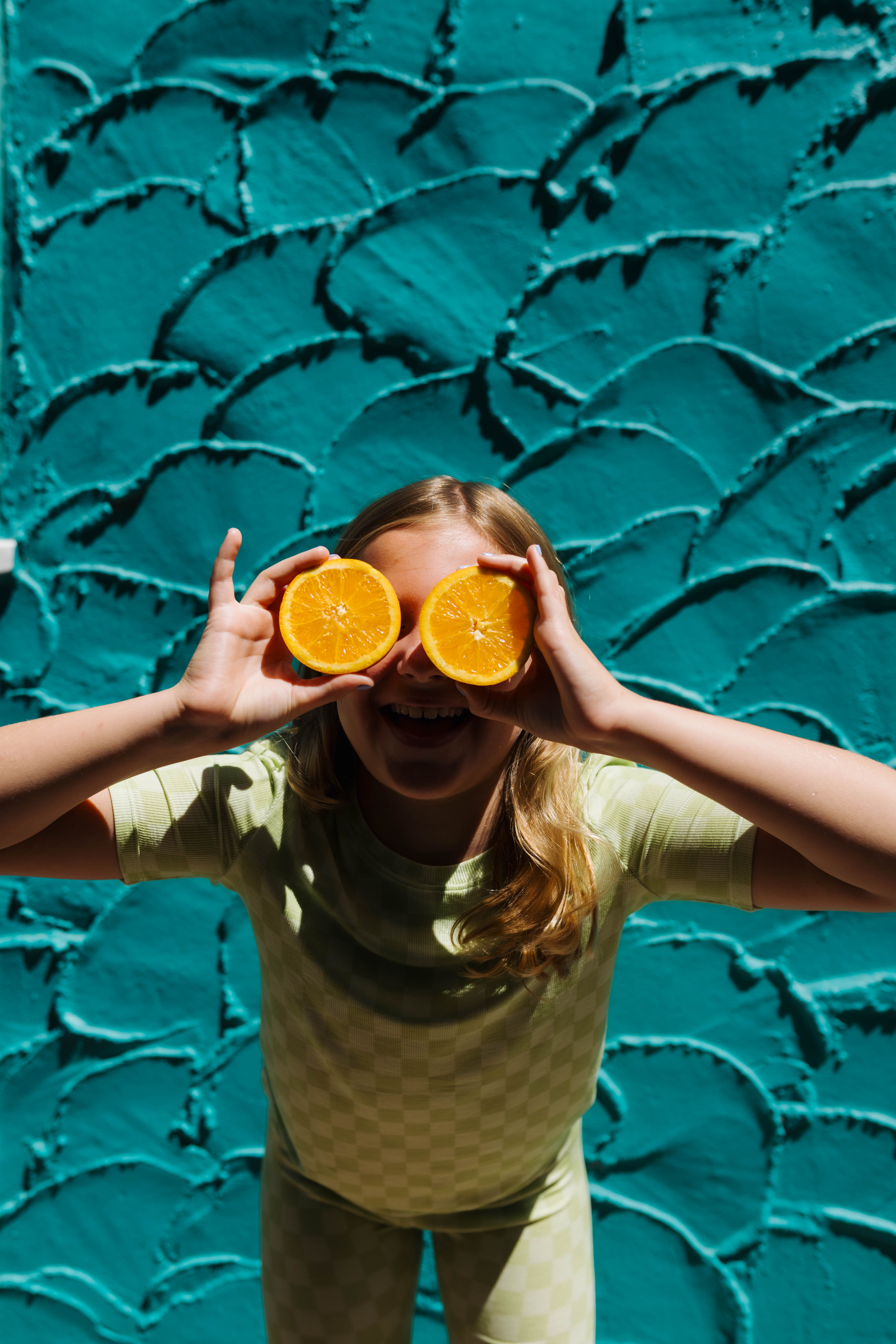A young girl with blonde hair wearing a light green check pjs holds two orange halves up to her eyes, posing playfully in front of a textured teal wall.