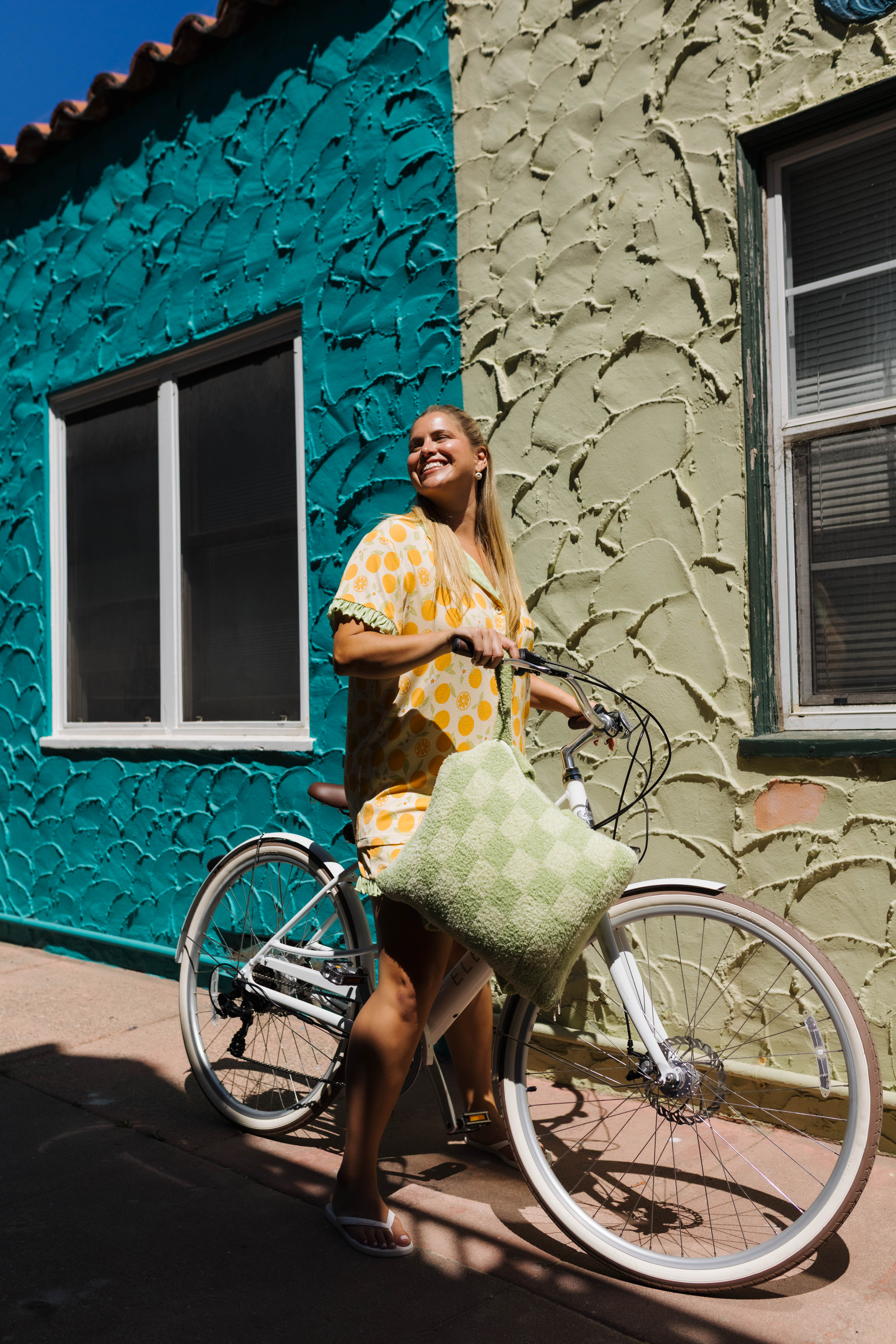 A woman smiling and riding a white bicycle outdoors next to a colorful textured building wall with a green check quillow