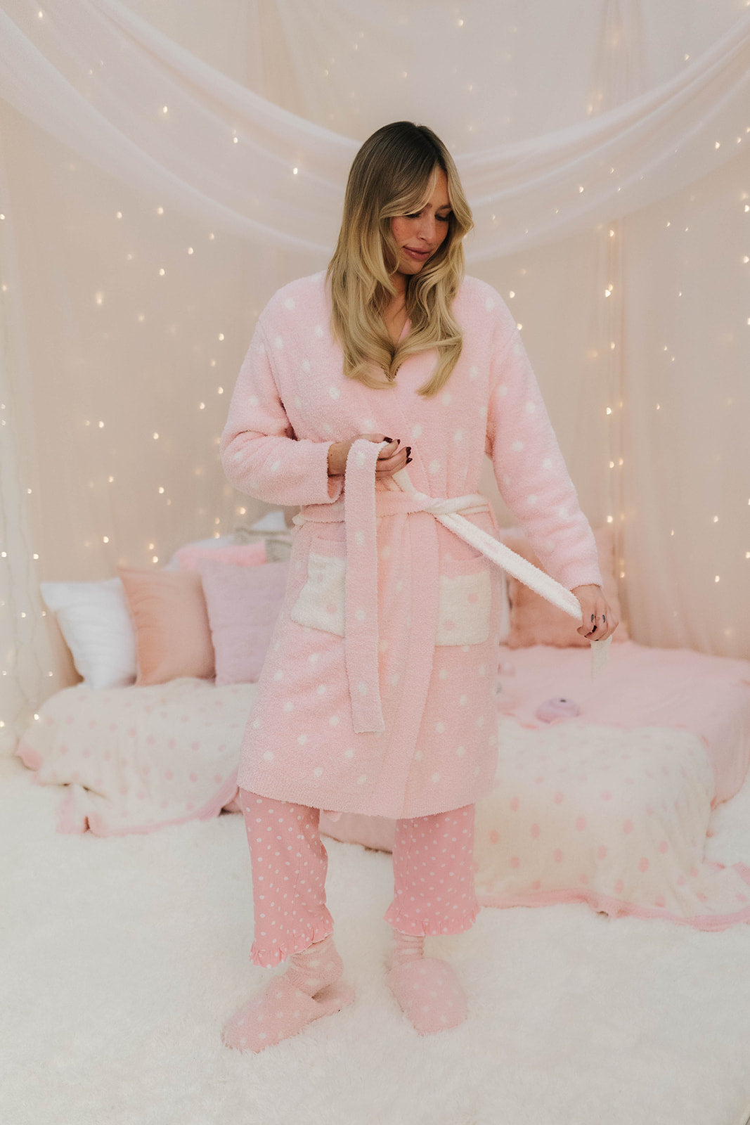 Woman in pink robe and pajama set by a bed with pink pillows and blanket.