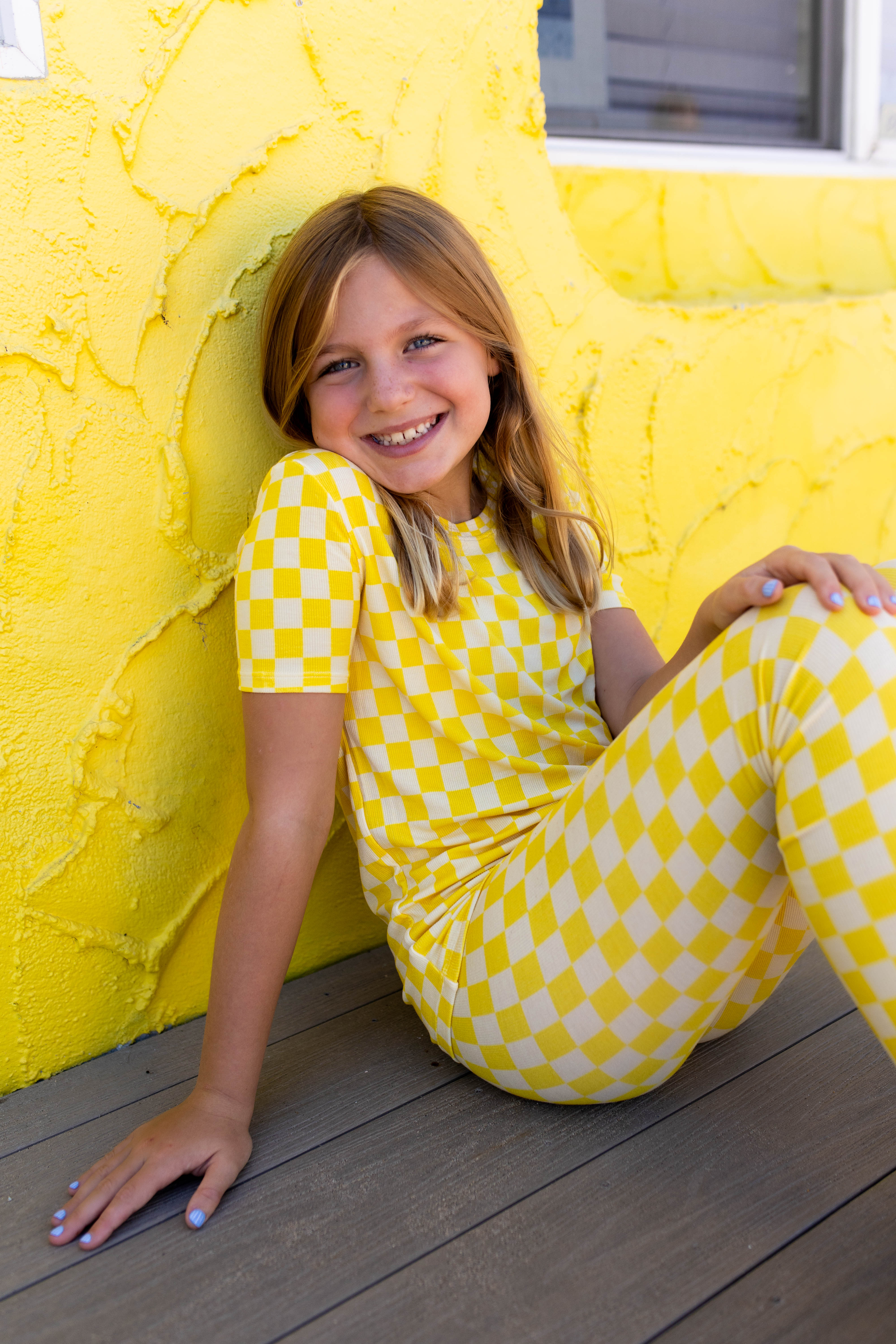 A young girl with blonde hair sits smiling on a wooden deck against a bright yellow stucco wall, wearing a matching yellow and checkered short-sleeve top pj set.