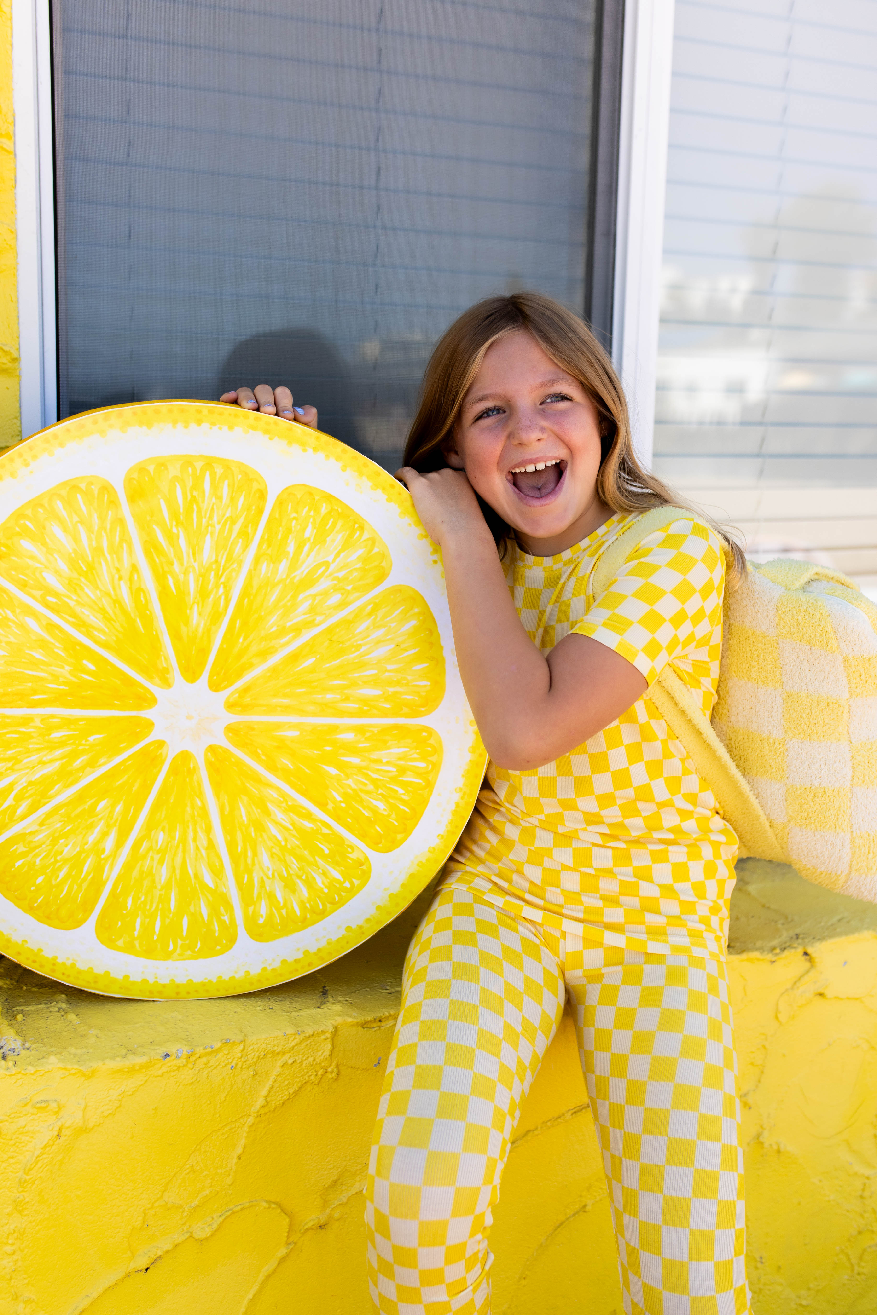 A young girl wearing a yellow and white checkered lemon-pattern pj set sits smiling next to a giant yellow lemon slice prop in front of a window.