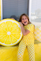 A young girl wearing a yellow and white checkered lemon-pattern pj set sits smiling next to a giant yellow lemon slice prop in front of a window.