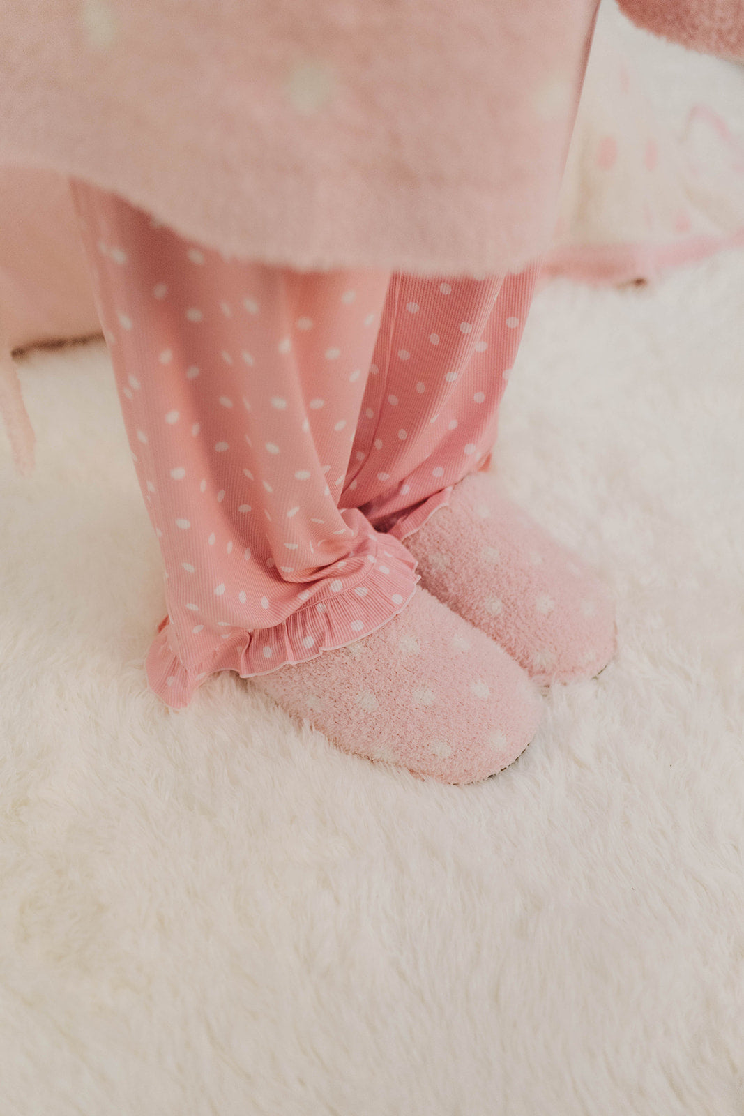 Pink polka dot pajama pants with ruffle trim worn by a person on a white surface.