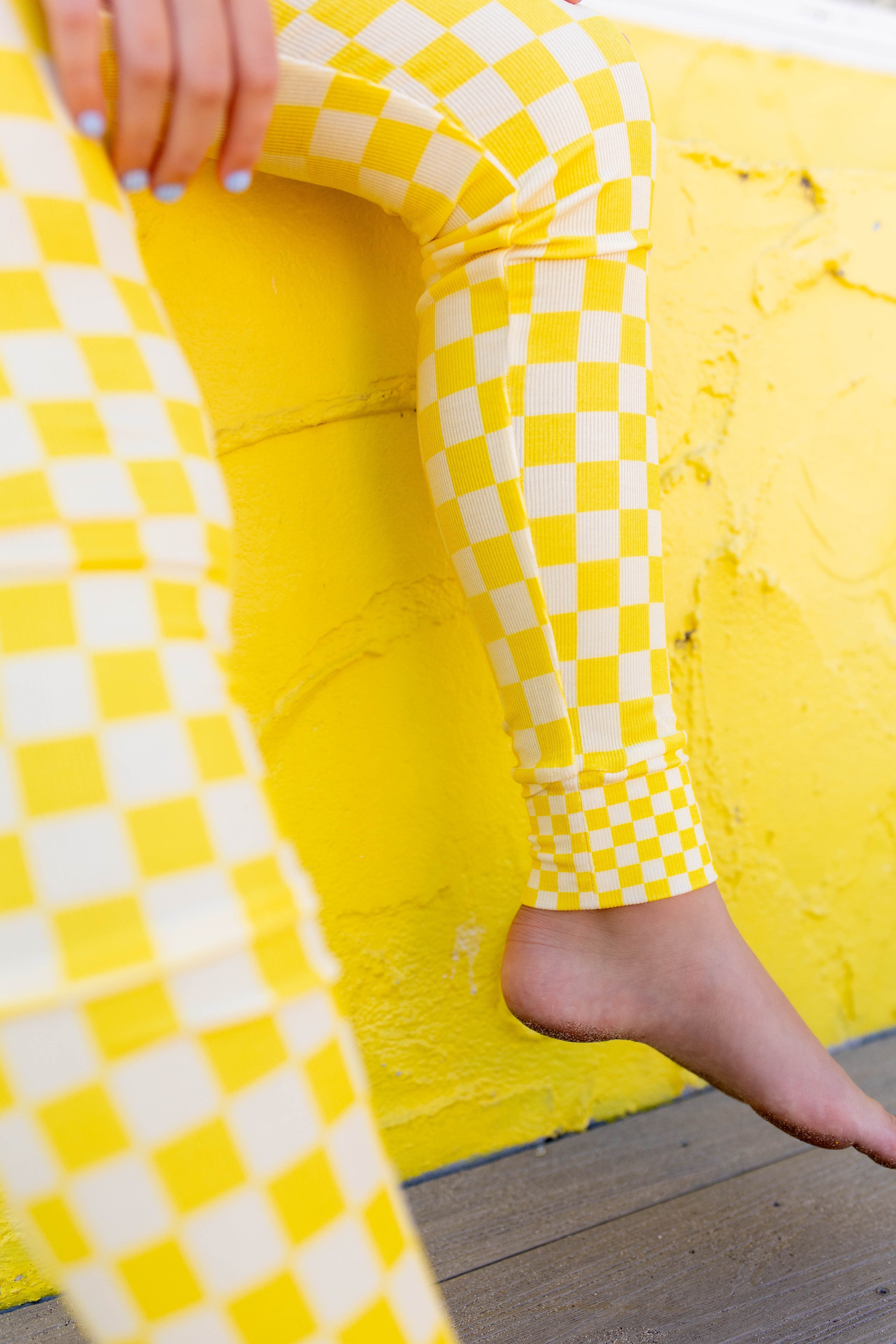 Close-up of a person wearing bright yellow and white checkerboard leggings, seated against a textured yellow wall with one bare foot visible.