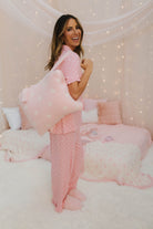 Woman in pink polka dot pajamas sitting on a bed with pink pillows and blankets.