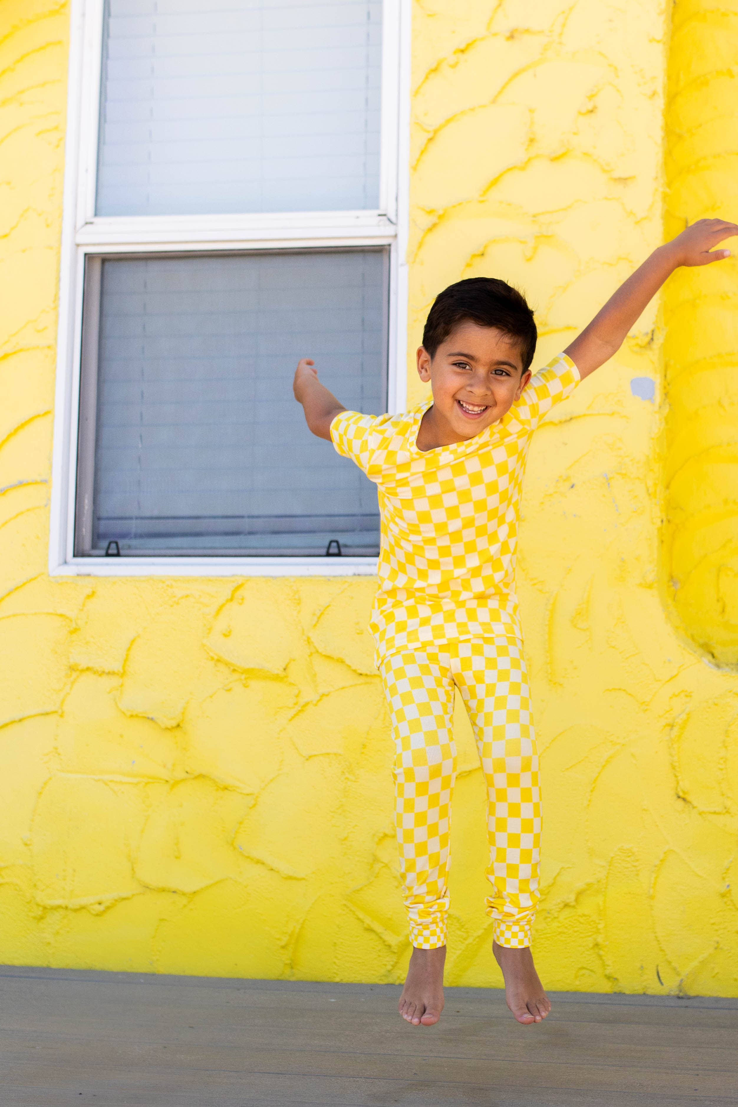 A young boy wearing a yellow checkered pajama set jumps in front of a bright yellow textured wall, with a window on the left.