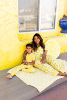 A woman and a young boy wearing coordinating pjs sit together on a light yellow blanket on a wooden deck in front of a bright yellow wall decorated with a lemon slice.