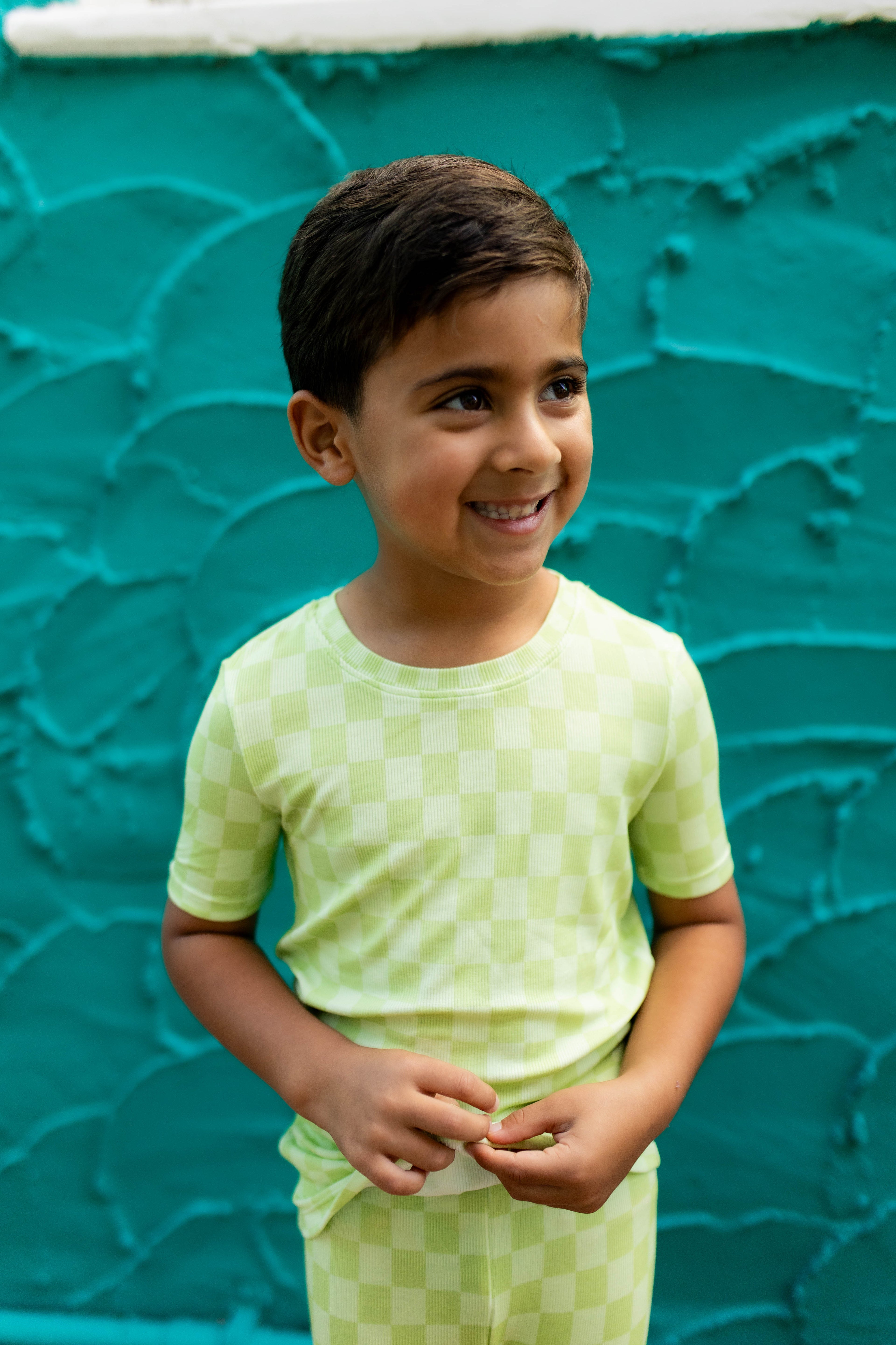 A young boy smiling and looking to the side, wearing a light green checkered short-sleeve t-shirt and matching pants, standing against a textured teal wall.