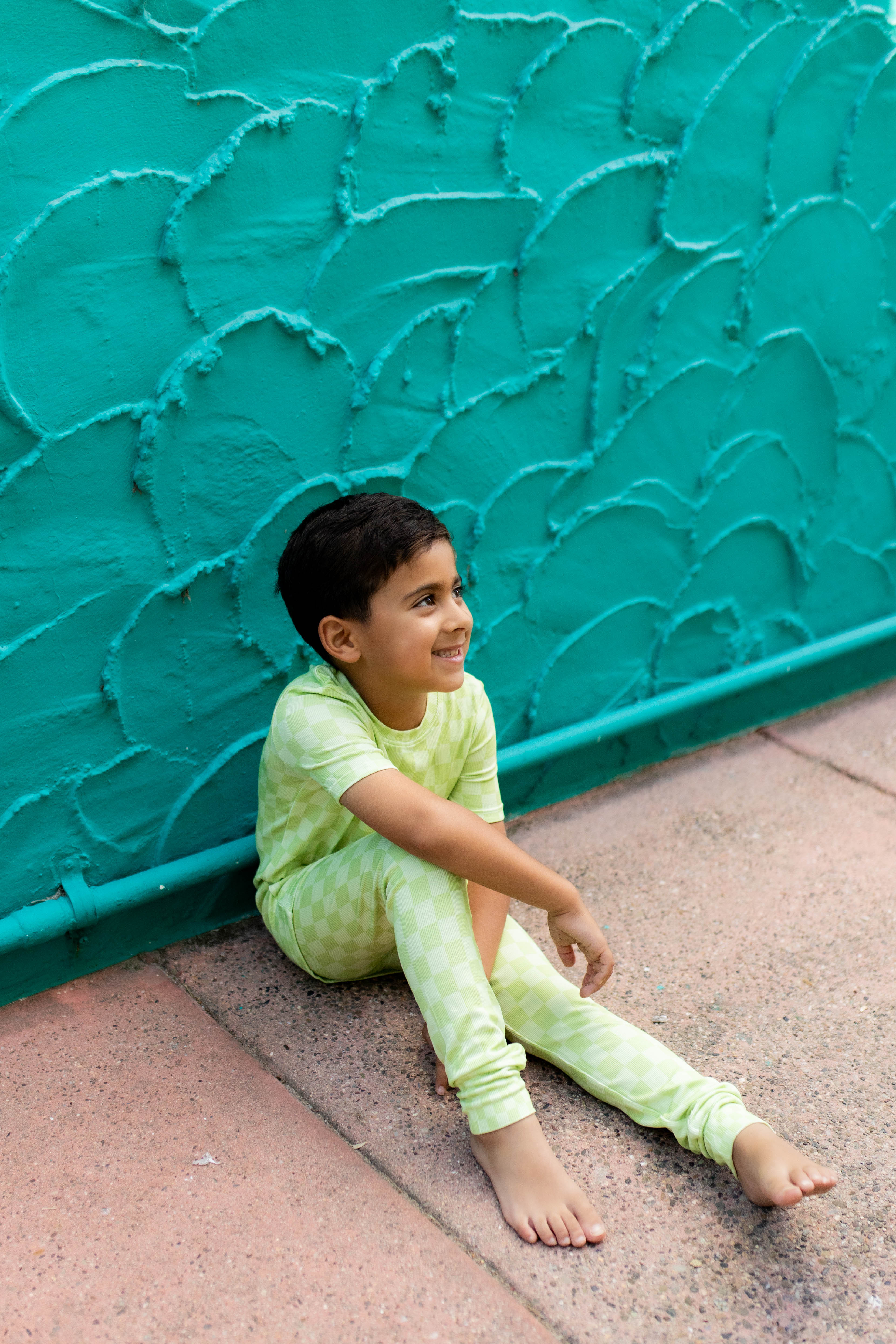A young boy wearing a light green check pajama set sits barefoot against a textured teal wall.