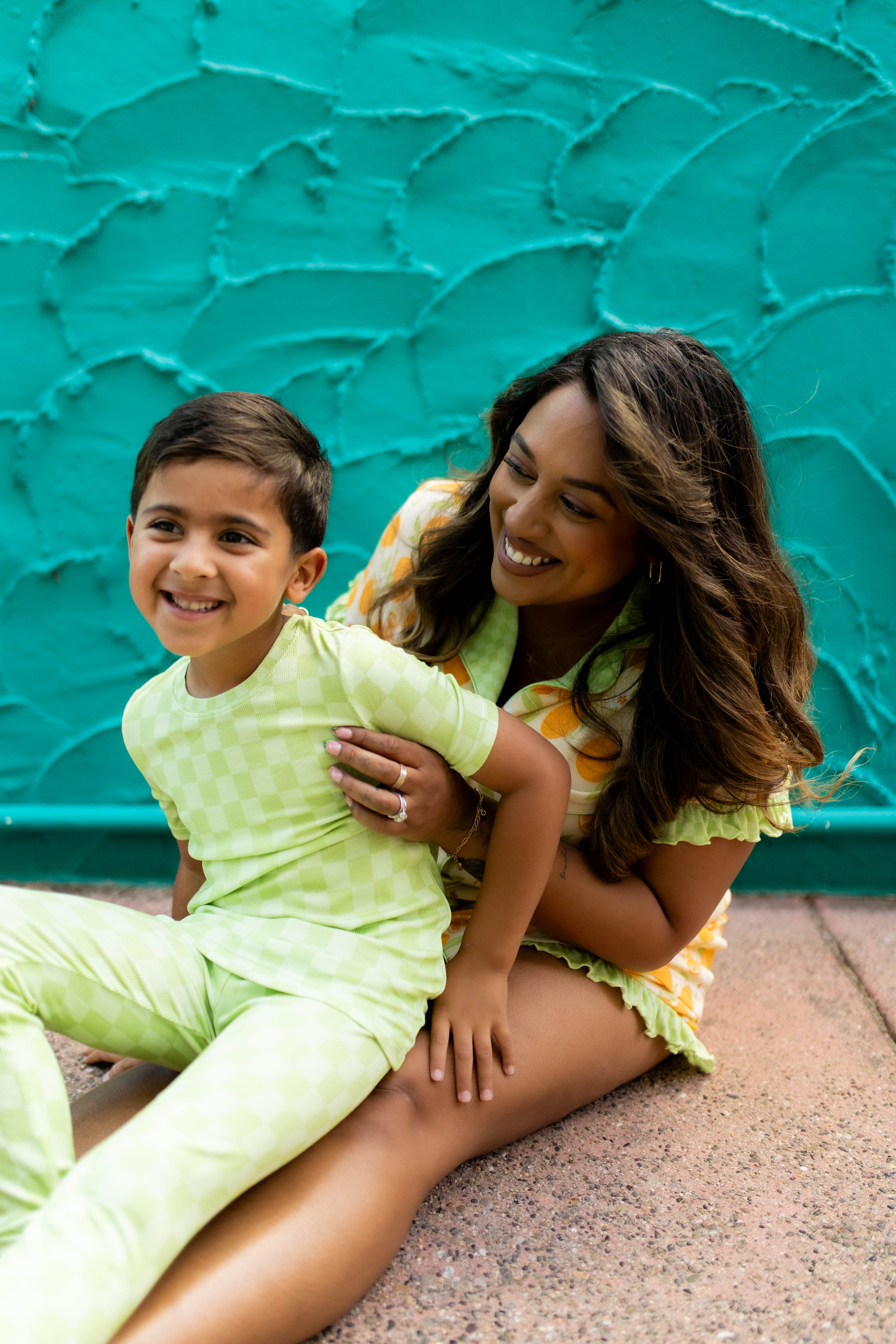 A woman and a young boy sitting together in front of a textured turquoise wall. The boy is wearing a light green checkered pajama set, and the woman is wearing a matching yellow and orange patterned pjs.