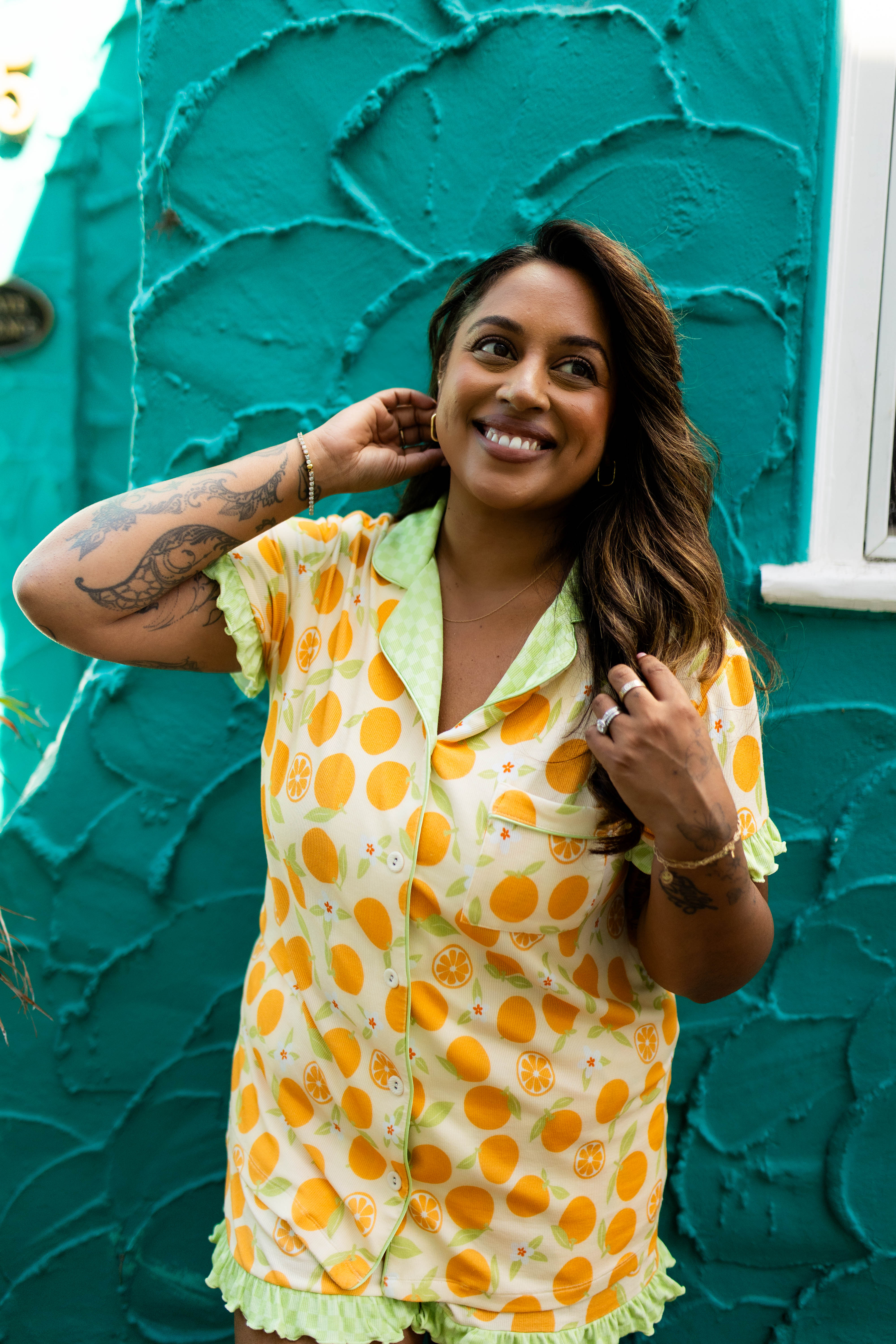 A woman smiling in a yellow and orange patterned short-sleeve pajama top with light green ruffled trim, standing in front of a textured teal wall.