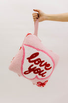 Pink pillow with 'love you' text held by a hand against a light background