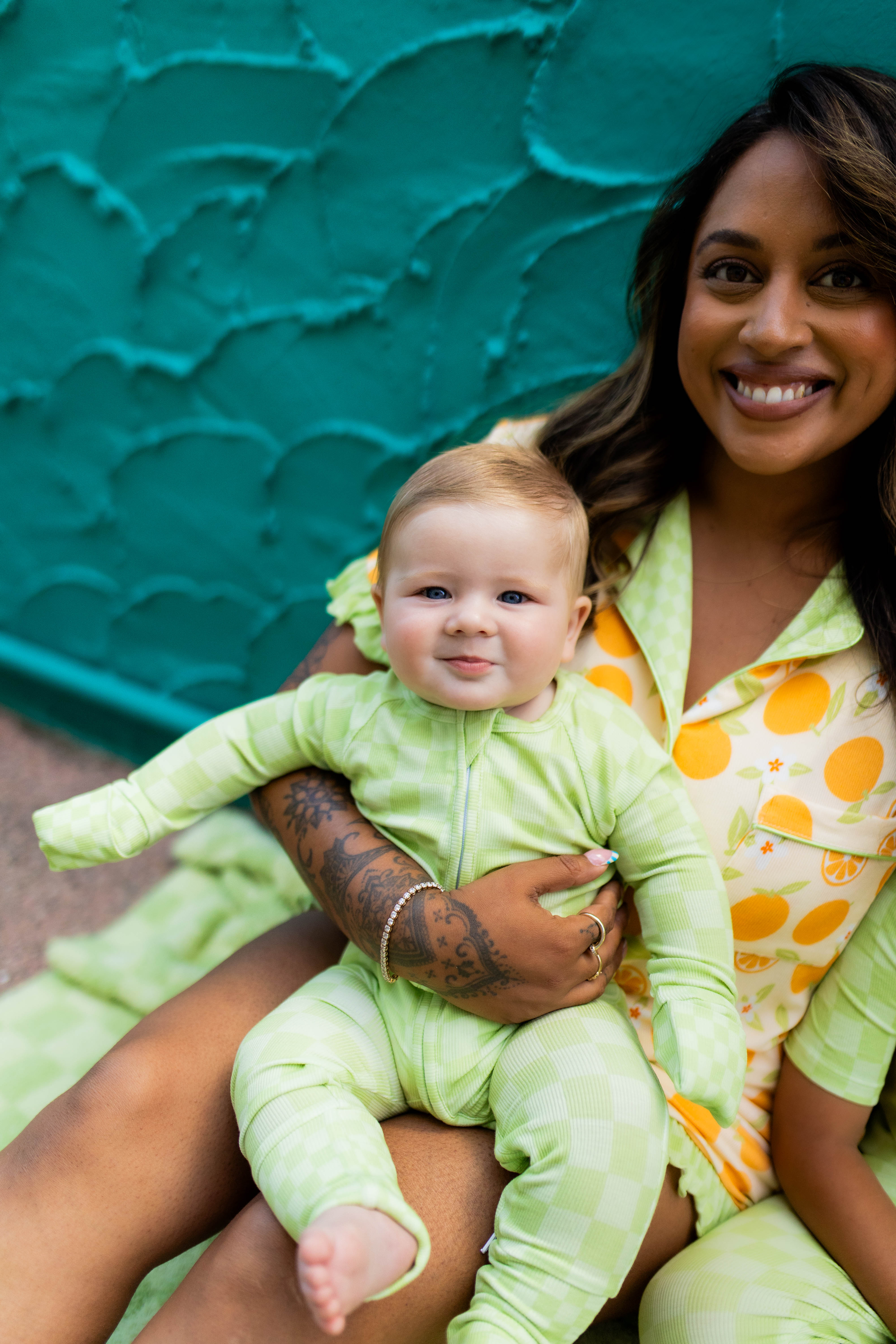 A smiling woman holding a baby wearing a light green footed pj, with the woman also wearing orange pjs, set against a textured teal wall.
