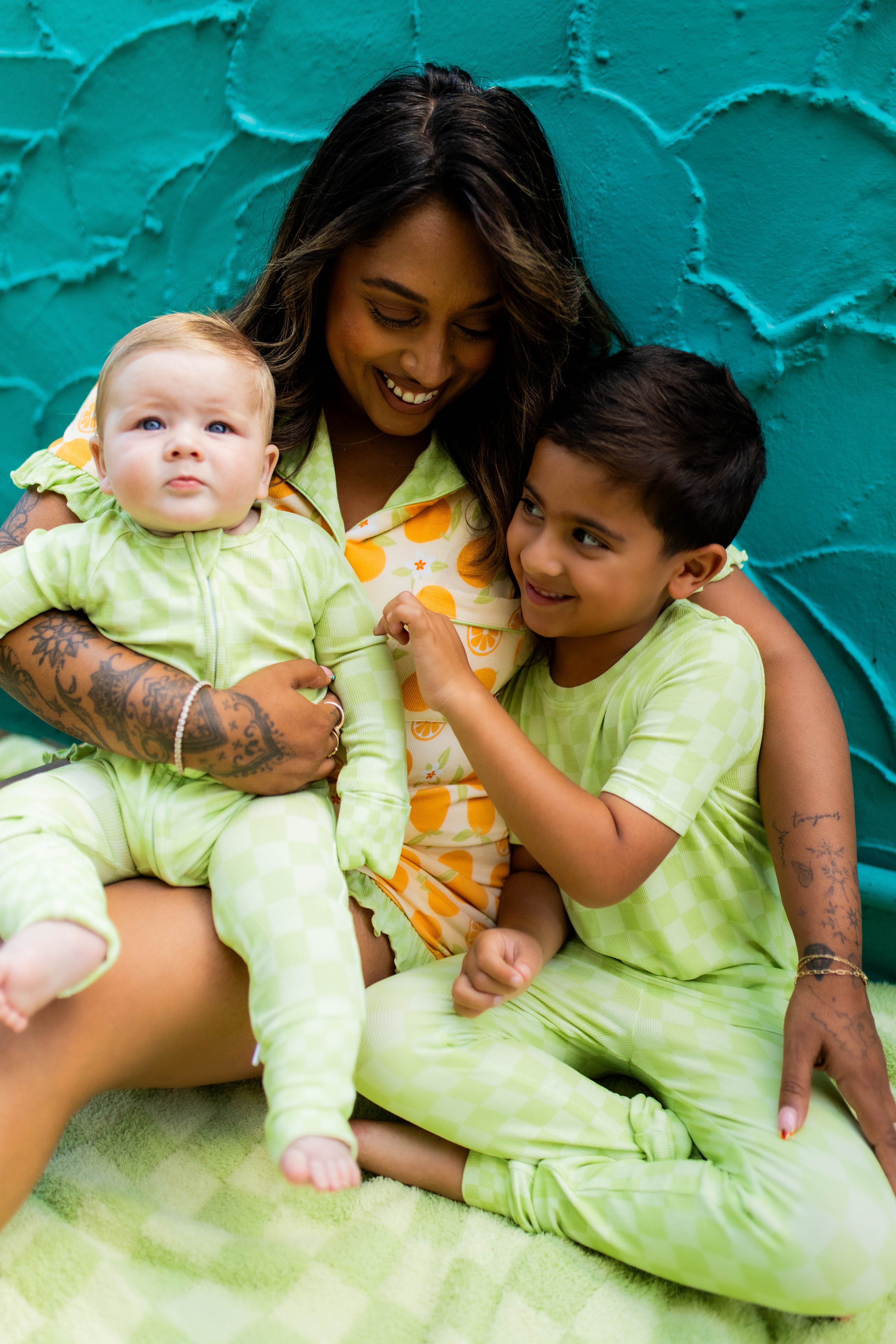 A woman and two children wearing matching light green pajamas sit together against a textured teal wall. The woman wears an orange patterned pajama top, while the children wear light green pajamas.