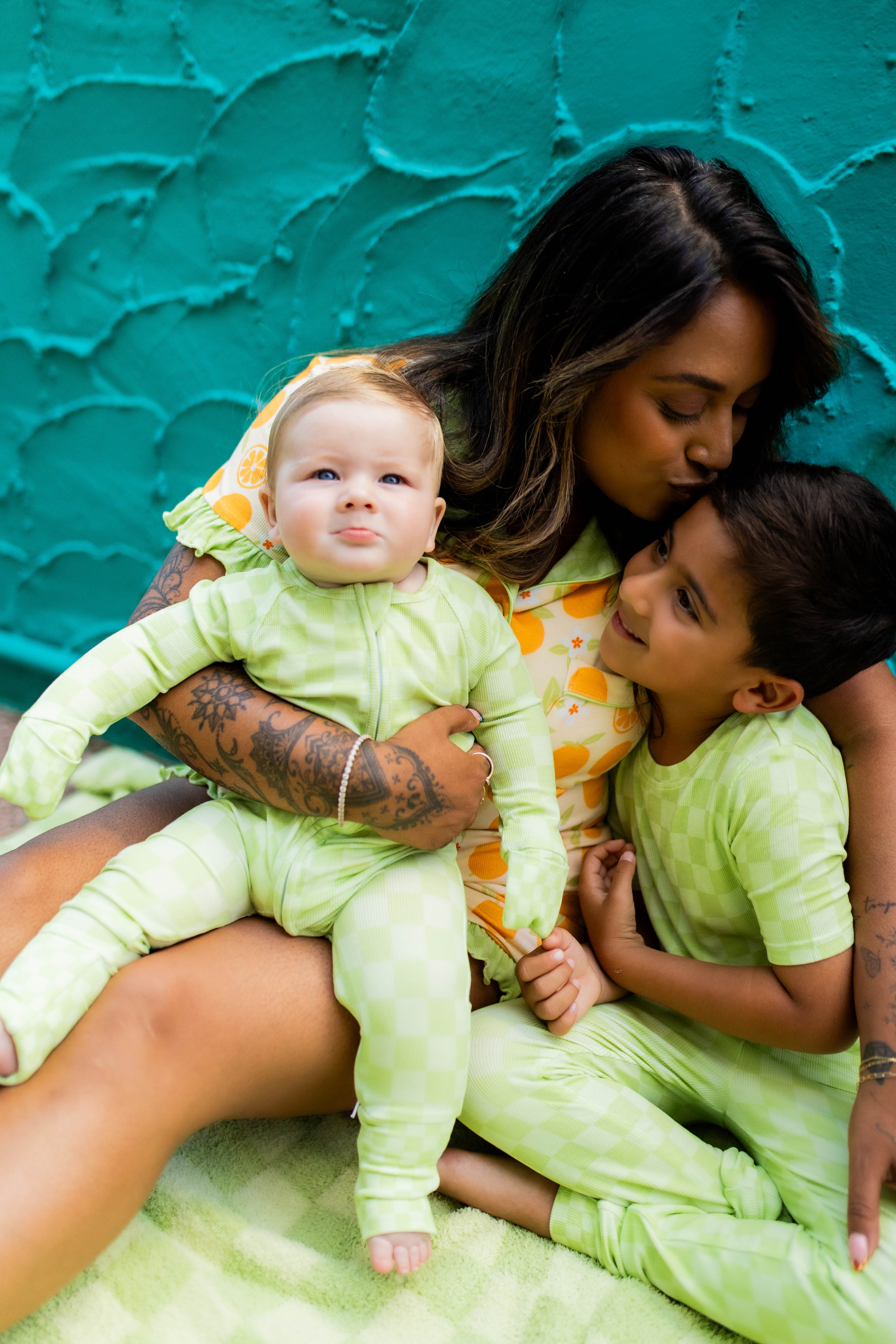 A mother and two children wearing matching light green patterned pajamas, sitting together against a textured teal wall.