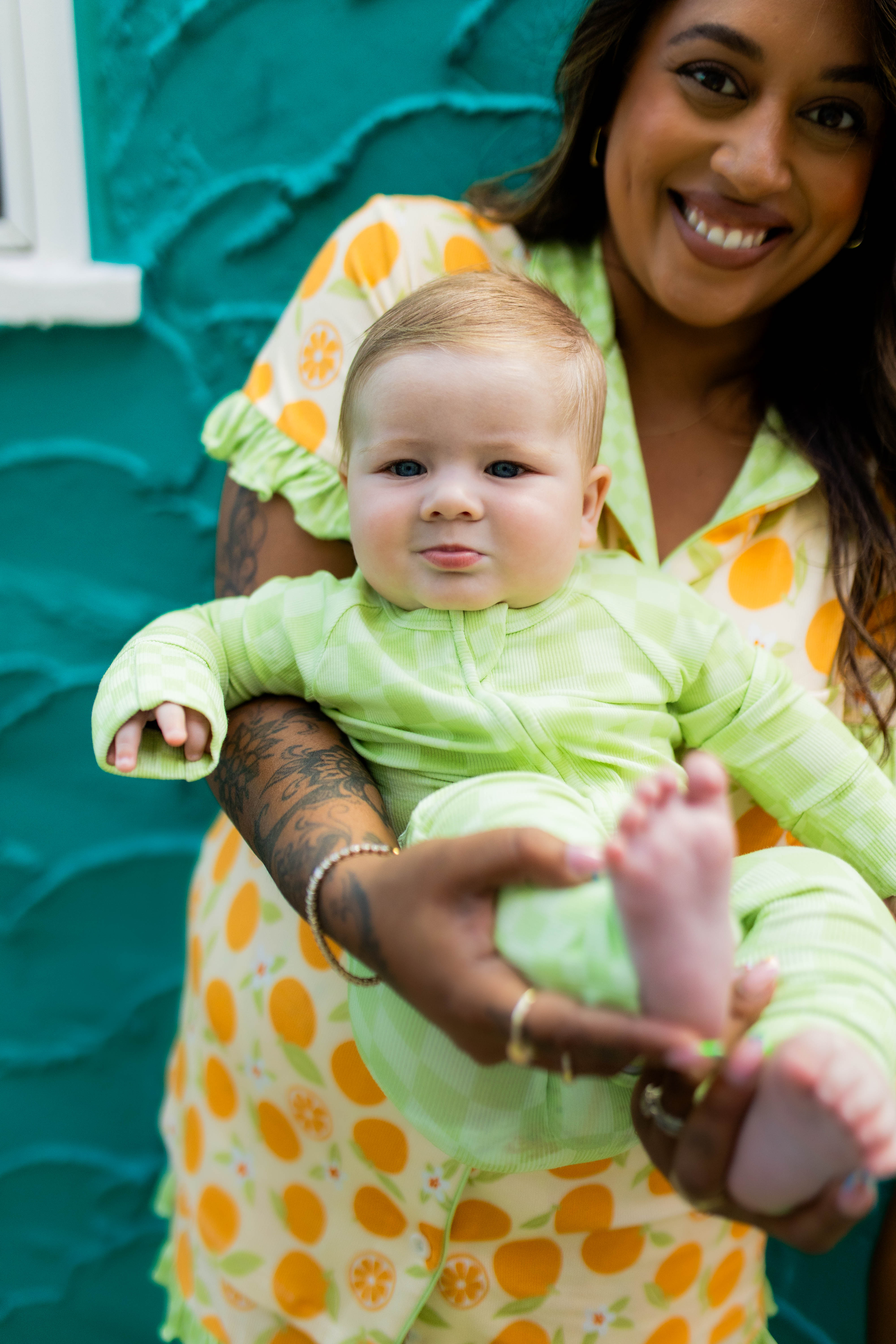 A smiling woman holds a baby wearing a light green long-sleeve outfit against a textured teal wall. The woman wears orange pjs.