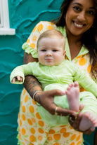 A smiling woman holds a baby wearing a light green long-sleeve outfit against a textured teal wall. The woman wears orange pjs.